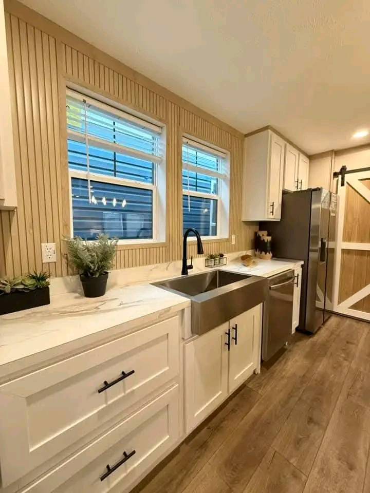 A modern kitchen with wood flooring, white cabinets, and a stainless steel farmhouse sink. Two windows above the sink add natural light, enhancing the cozy ambiance.