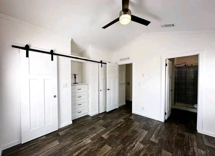 Minimalist room with white walls, dark wood floor, black ceiling fan, sliding barn doors, and an adjacent bathroom with a patterned shower curtain.