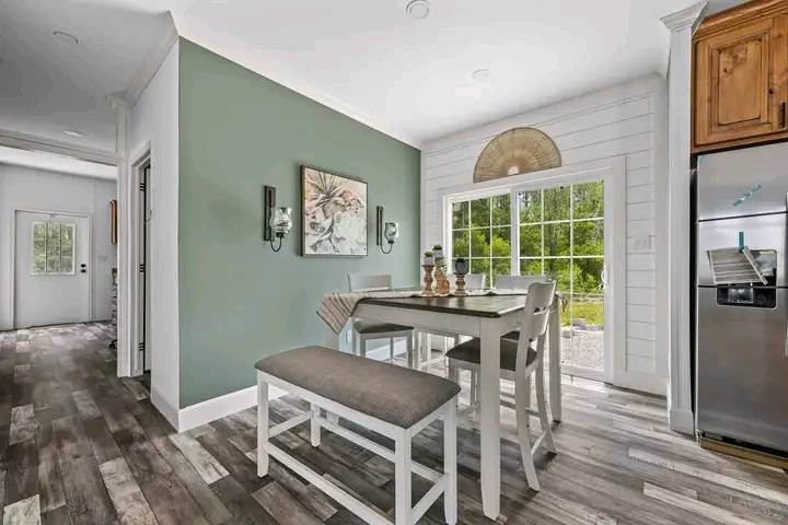 Bright dining area with a wooden table, cushioned bench, and chairs. A green accent wall with art, sconces, and large glass doors open to a garden.