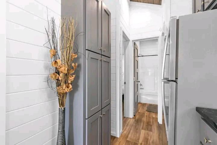 Narrow hallway in a modern home with white shiplap walls, gray cabinets, brown hardwood floors, and a tall vase with dried orange flowers.