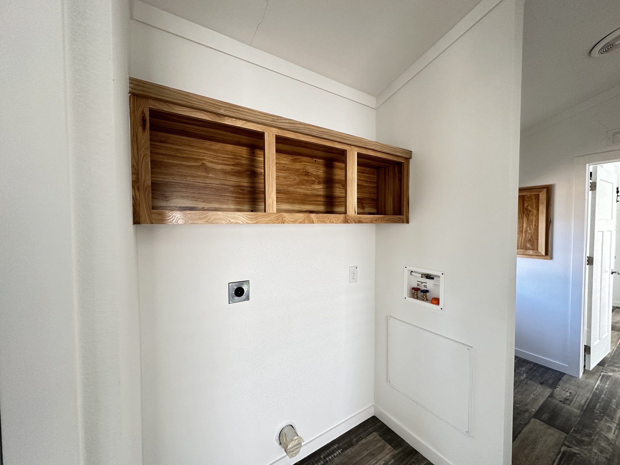 Laundry room with white walls featuring natural wood open shelving. The floor is dark wood, and a utility hookup area is visible. The setting is modern and minimalistic.