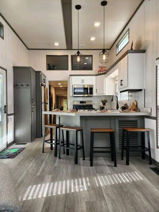 Modern kitchen featuring a gray and white color scheme, with a central island, bar stools, hanging pendant lights, stainless steel appliances, and wood flooring.