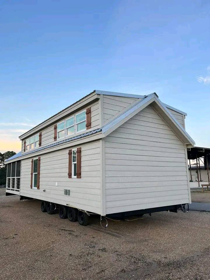 A beige tiny house on wheels sits on a gravel surface under a clear blue sky. It features multiple windows with wooden shutters, conveying a sense of mobility.