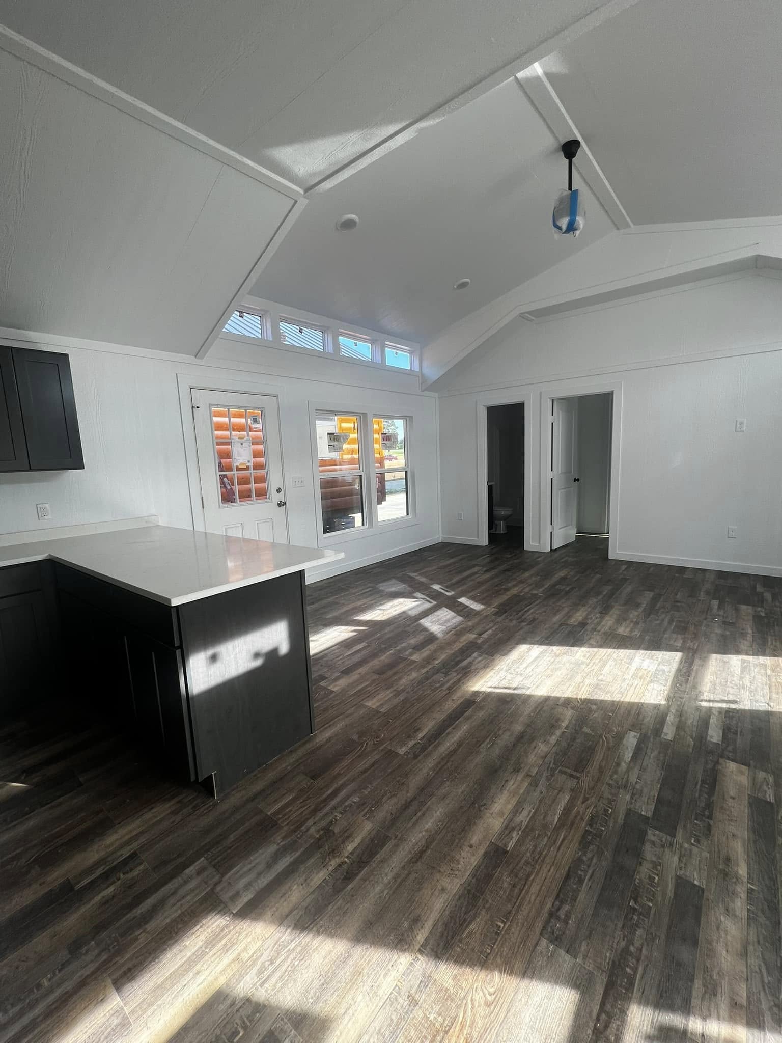 A spacious, sunlit room with vaulted ceilings, wood floors, and white walls. French doors lead outside, and a dark kitchen counter is visible.