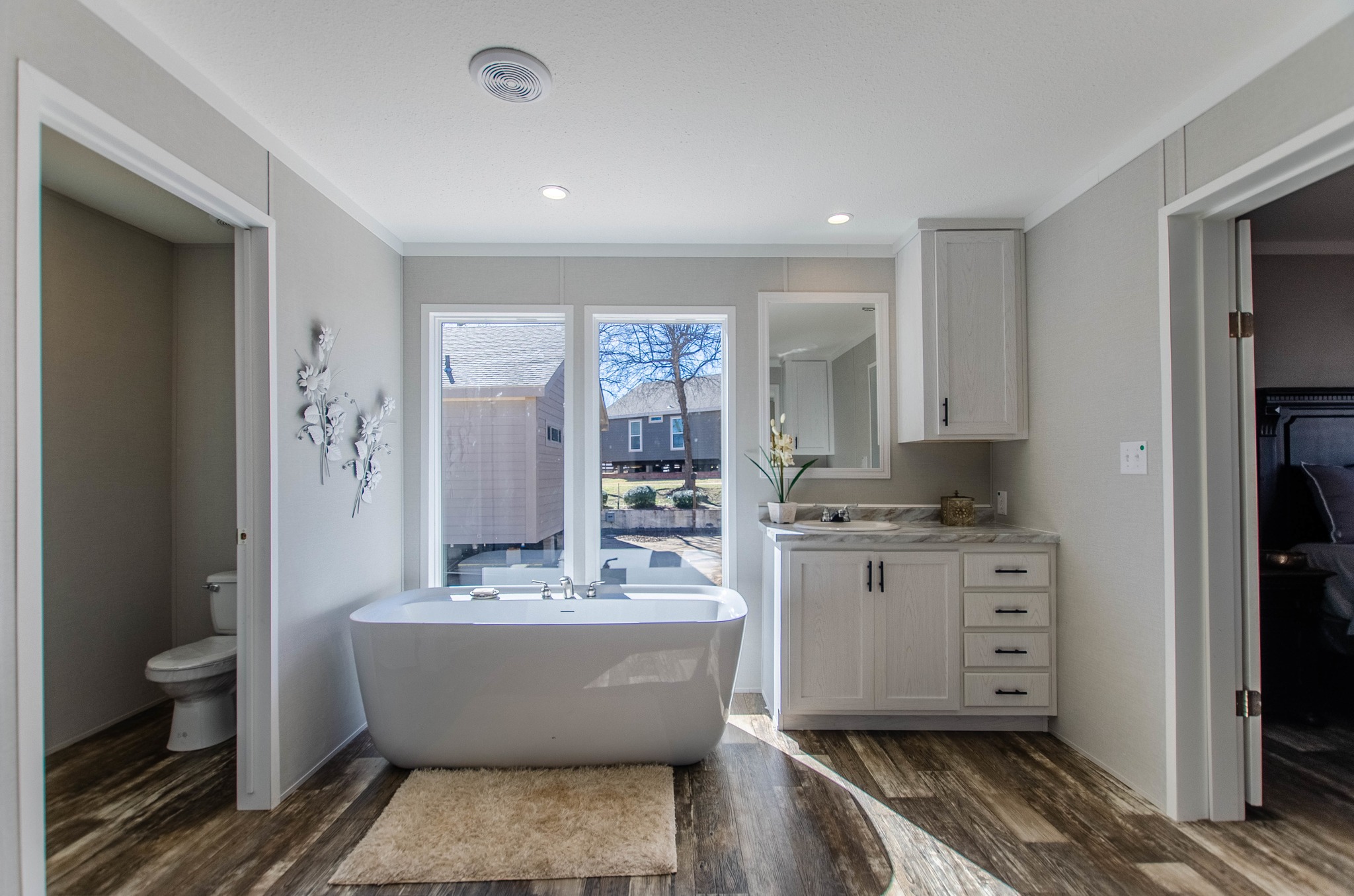 Modern bathroom with freestanding bathtub under large windows, wooden floor, white cabinetry, and decor creating a serene and airy atmosphere.