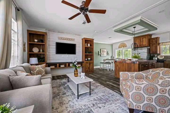 A cozy living room with a modern design, featuring a neutral-toned sofa and patterned armchair. A ceiling fan hangs overhead. The area seamlessly connects to a kitchen with wooden cabinets and a stylish island. A wall-mounted TV is flanked by built-in shelves. The atmosphere is inviting and elegant.