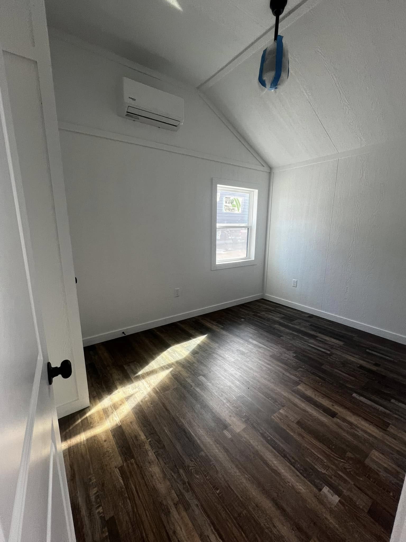 A bright, empty room with white walls, a sloped ceiling, and a wood floor. Sunlight streams through a window, illuminating the space. A mini-split AC unit is mounted above the window. The room feels clean and serene.