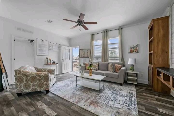 Modern living room with gray sofa, patterned armchair, and a wooden bookshelf. A ceiling fan hangs above a coffee table on a patterned rug, exuding a cozy, inviting atmosphere.