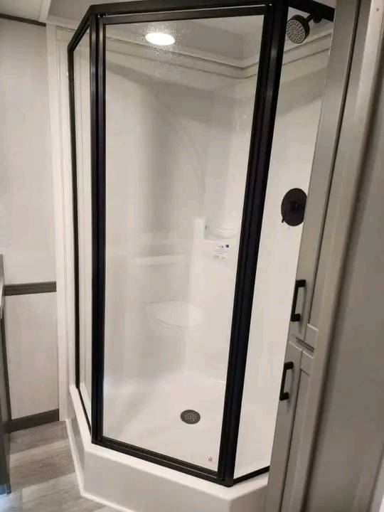 Compact corner shower with clear glass panels and a black frame. Inside, a small white bench and a showerhead are visible, creating a clean and modern feel.