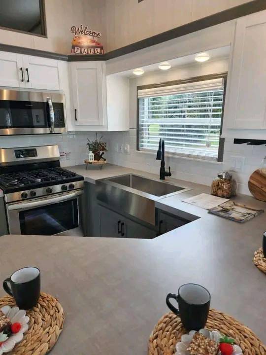Modern kitchen with stainless steel appliances, white cabinets, and a sink under a window with blinds. Counter has mugs on woven mats, exuding a cozy vibe.
