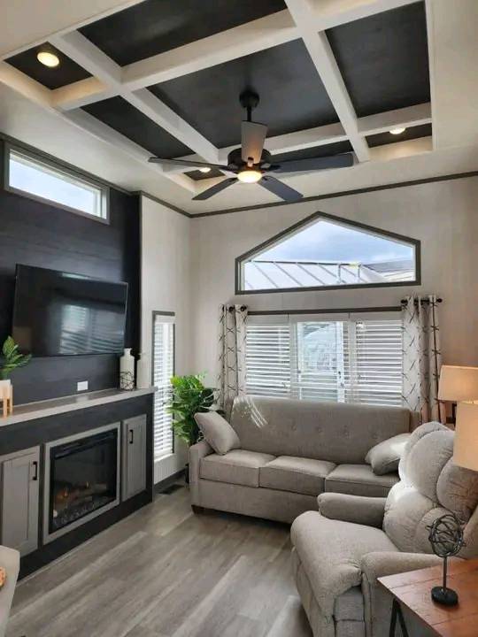 Modern living room with a coffered ceiling, ceiling fan, and a large TV above a sleek fireplace. Cozy grey sofas, plants, and bright natural light fill the space.