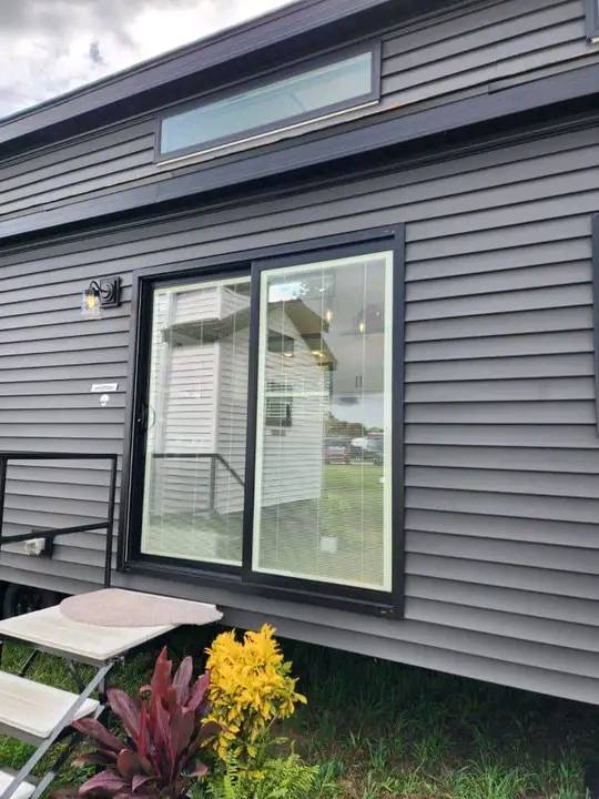 A modern tiny house with gray siding and a large glass sliding door. A small deck with steps leads up to the entrance. A potted red and yellow plant is in the foreground, adding a touch of nature.