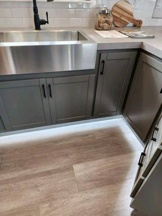 Modern kitchen with dark cabinets, stainless steel sink, and black faucet. Under-cabinet lighting highlights wood flooring. Cozy and clean ambiance.