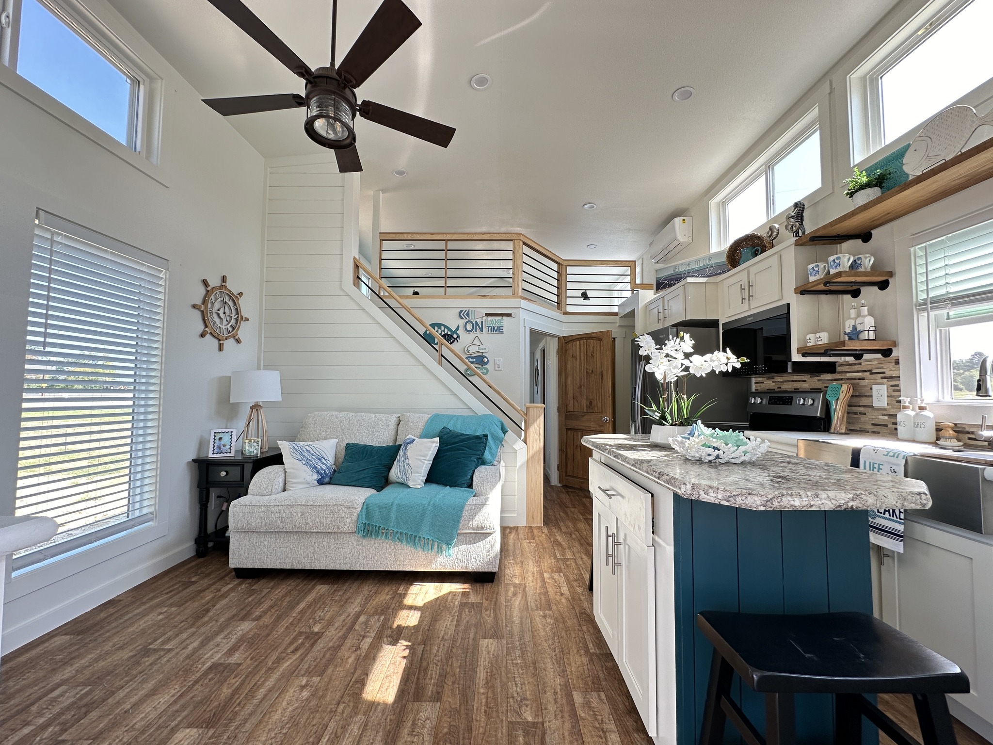 Bright, cozy living room with white walls, wooden floors, and a ceiling fan. A comfy sofa with teal accents faces a modern kitchen and stairs.