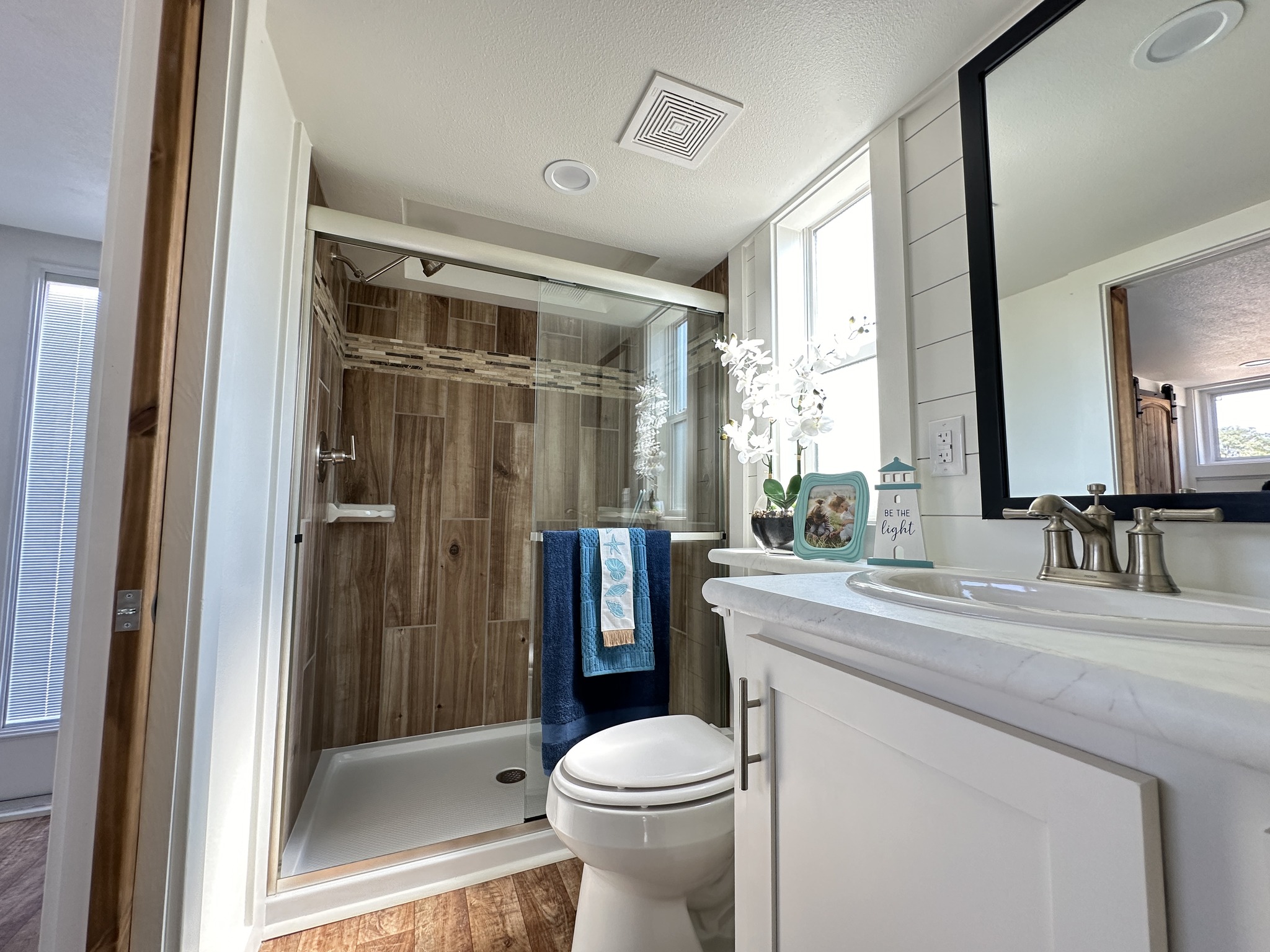 Bright and modern bathroom with wood-paneled shower, glass door, white vanity, decorative items, and a large mirror, creating a fresh atmosphere.