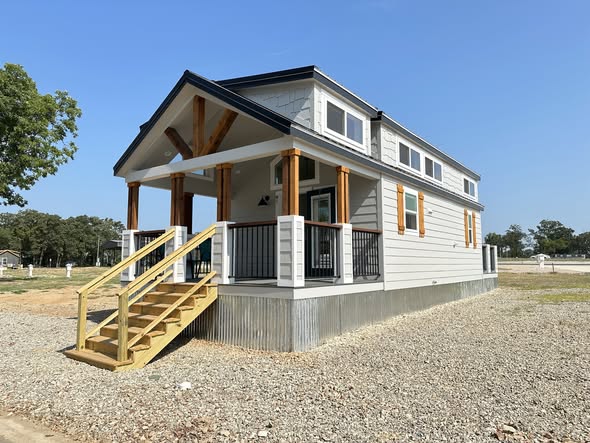 A modern tiny house with a raised porch and wooden stairs, set on gravel. It features white siding, large windows, and exudes a cozy, welcoming vibe.