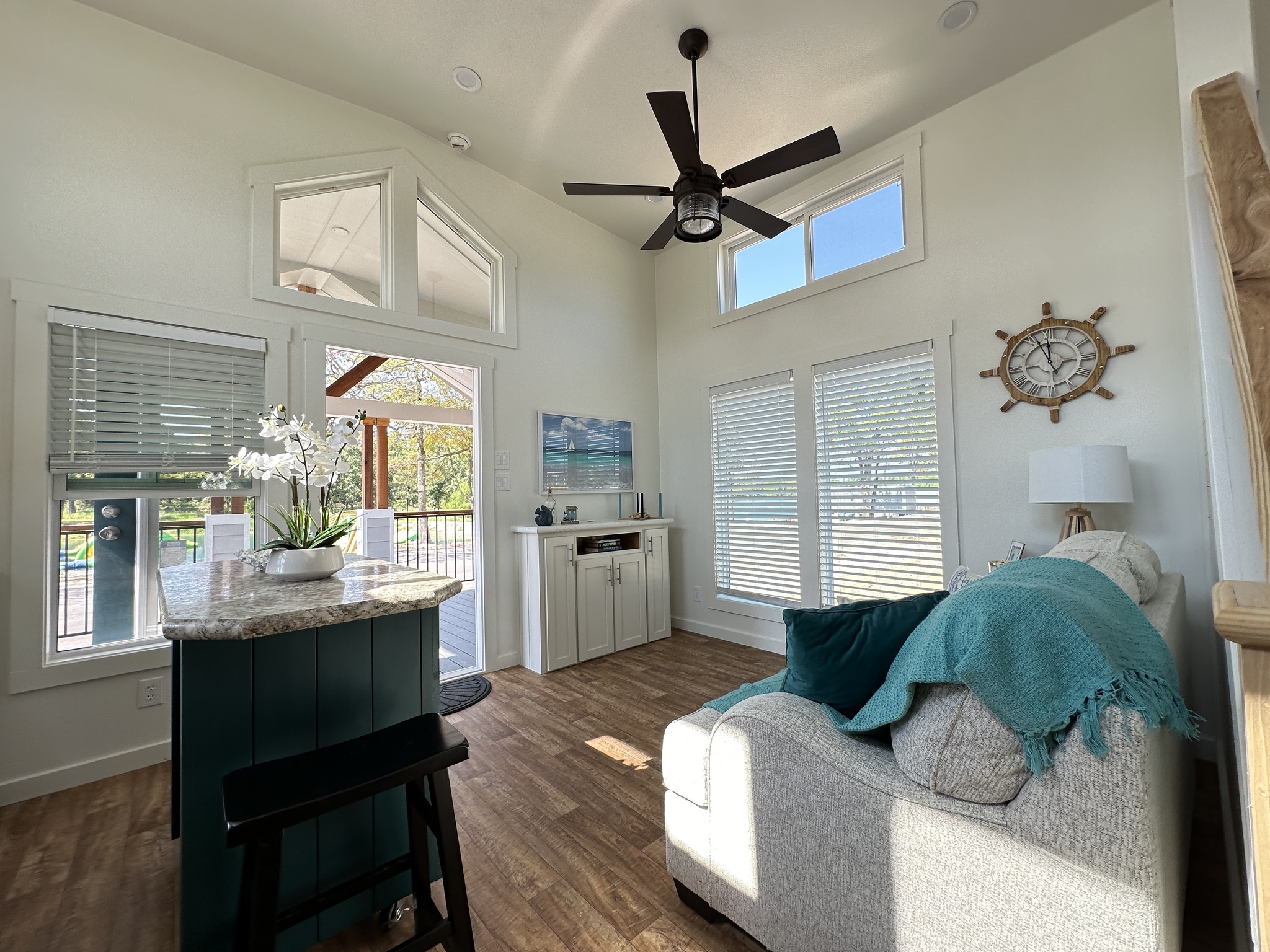 Bright, airy living room with tall ceilings, a ceiling fan, and large windows. Decor includes a helm clock, turquoise blanket, and orchid on a kitchen island.