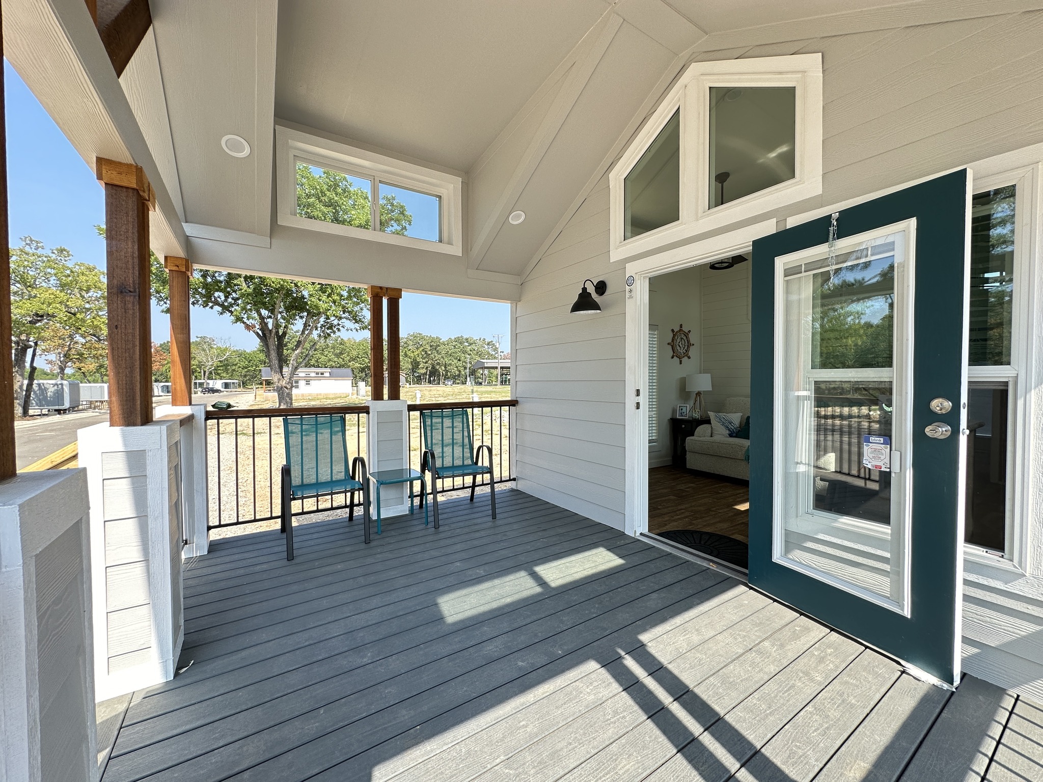 A cozy porch with two teal chairs, wooden beams, and an open door leading to a bright living room. Sun casts soft shadows, creating a welcoming feel.