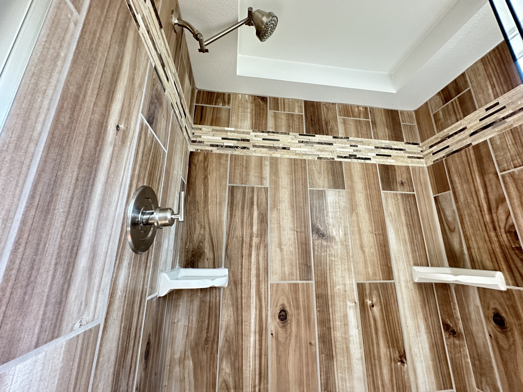 Wood-patterned bathroom shower with beige and brown tiles, accented by a mosaic border. Silver fixtures include a showerhead and control knob.