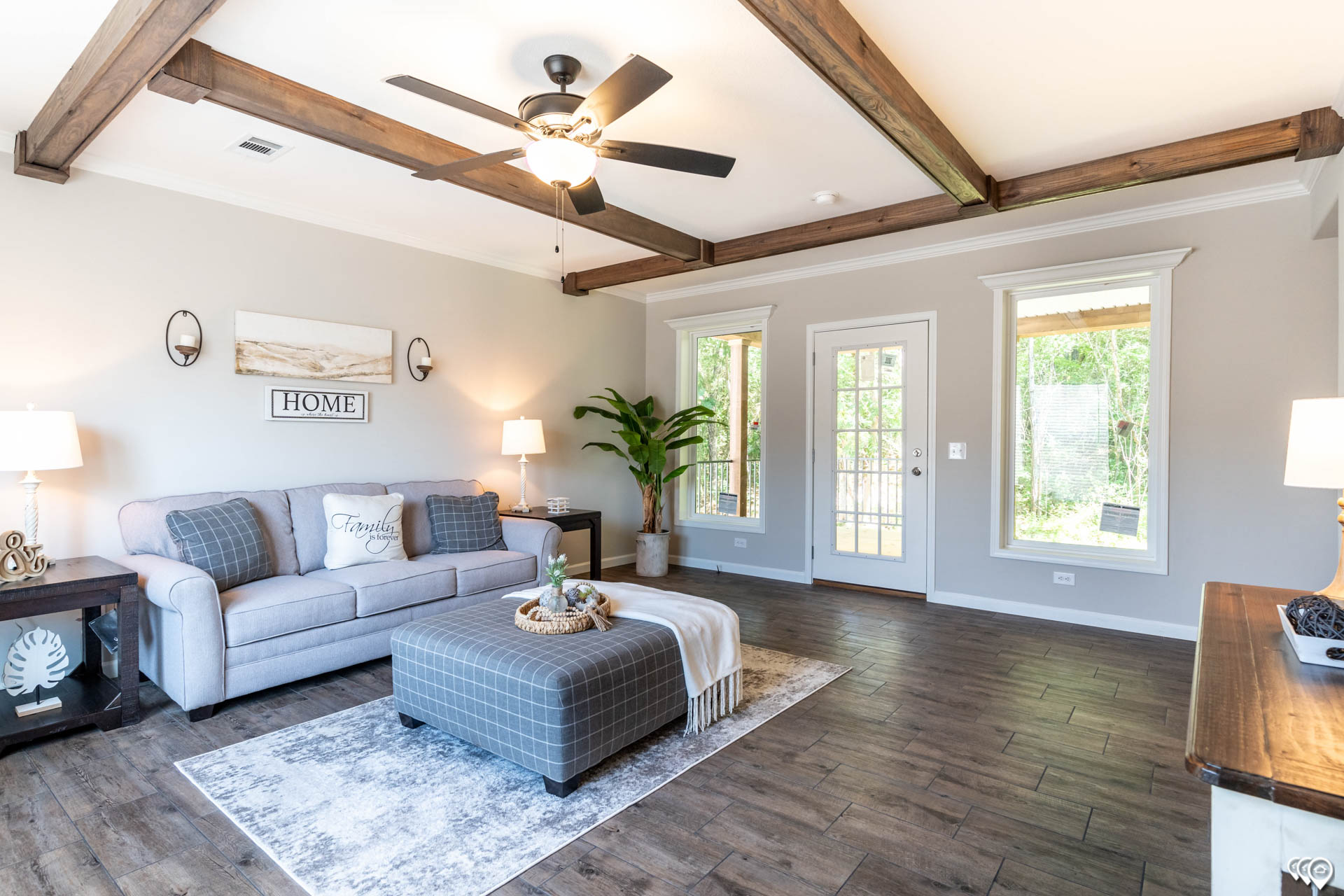 Cozy living room with light gray sofa, patterned ottoman, and soft lighting. Wooden beams on the ceiling and large windows create an inviting, airy space.