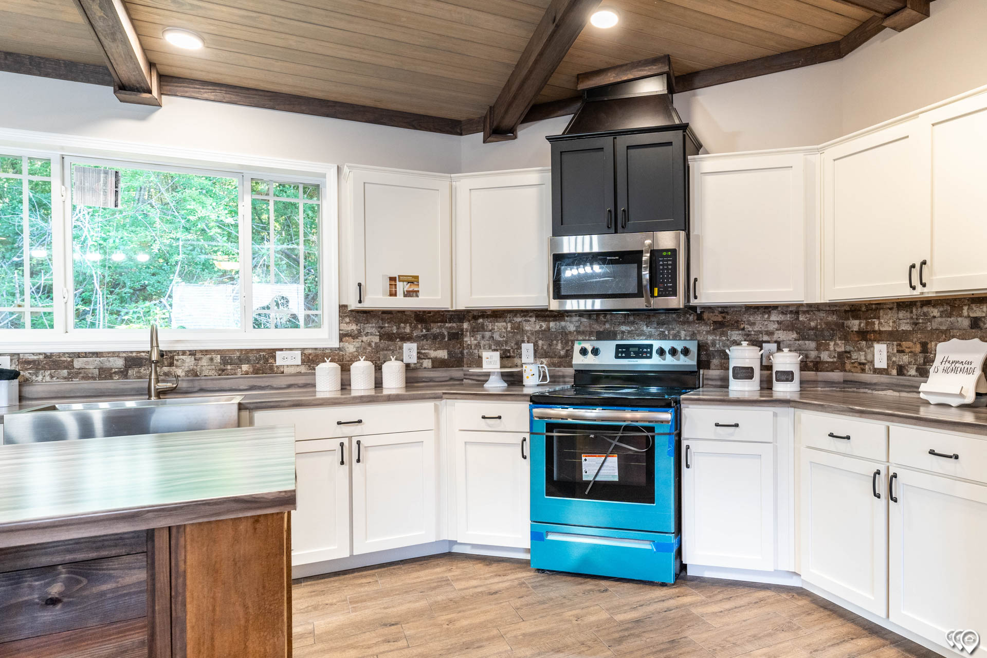 Modern kitchen with vaulted wooden ceiling, featuring a bright blue oven, black and white cabinets, and a large window overlooking greenery. Cozy and inviting.