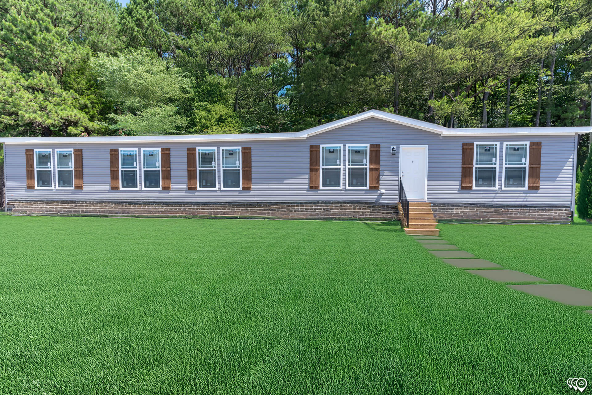 A single-story, gray manufactured home with brown shutters, sits on a large, vibrant green lawn. A path leads to the front door, surrounded by lush trees.