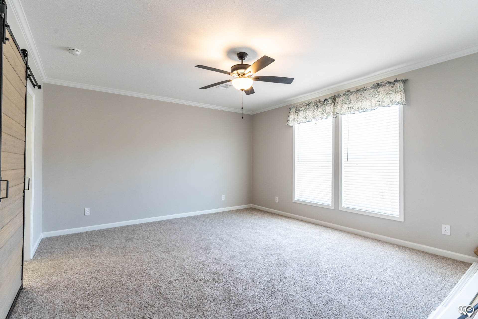 Empty room with beige carpet and light gray walls. A ceiling fan with a light is centered. Two windows with blinds allow natural light in.