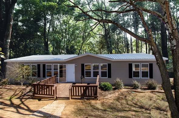Single-story gray house with black shutters, nestled among tall trees. A wooden ramp leads to the entrance, surrounded by soft landscaping on a sunny day.