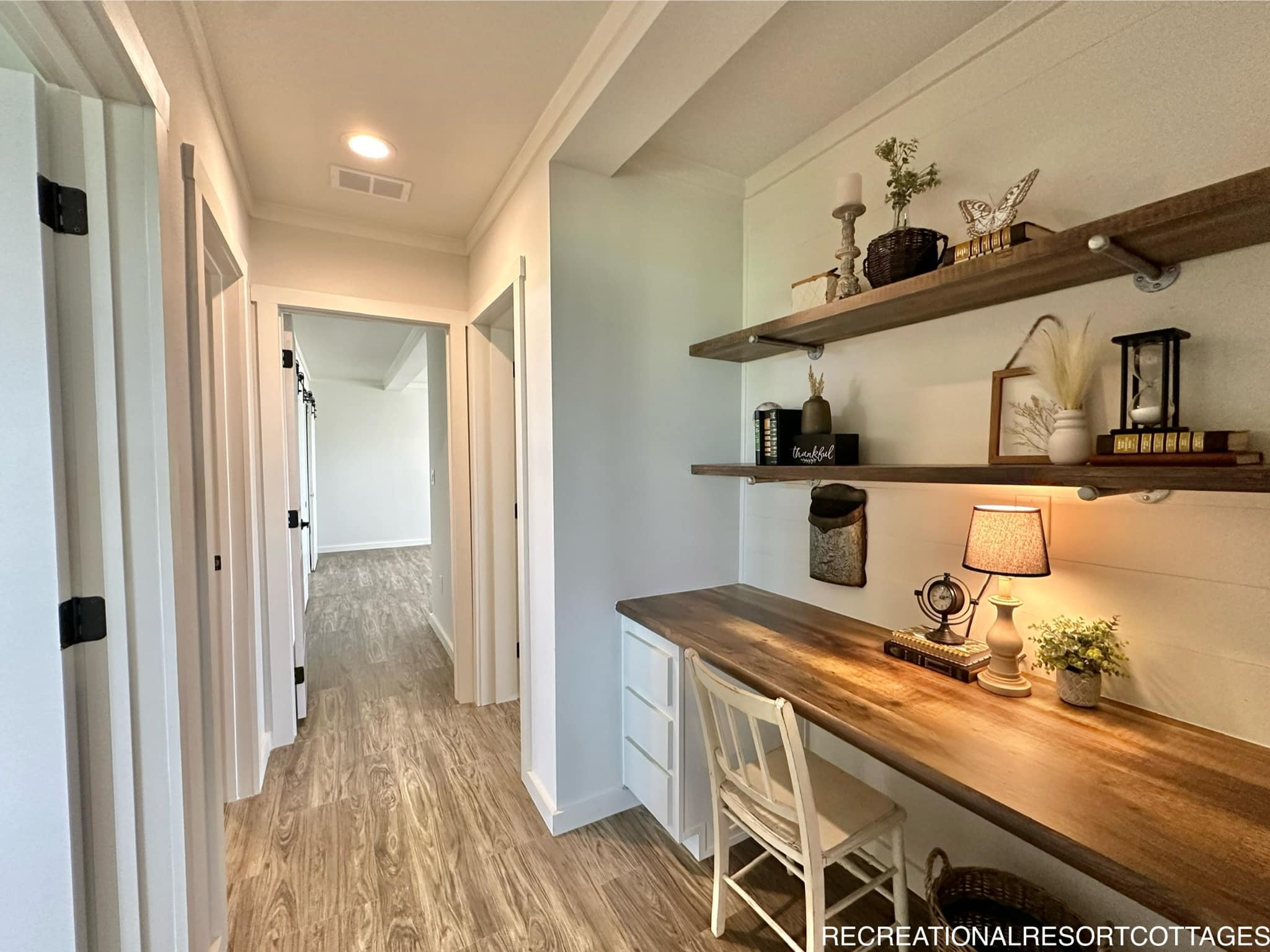 A cozy hallway with light wood flooring, a built-in desk, and shelves on the right adorned with plants and decor. A chair and lit lamp add warmth.