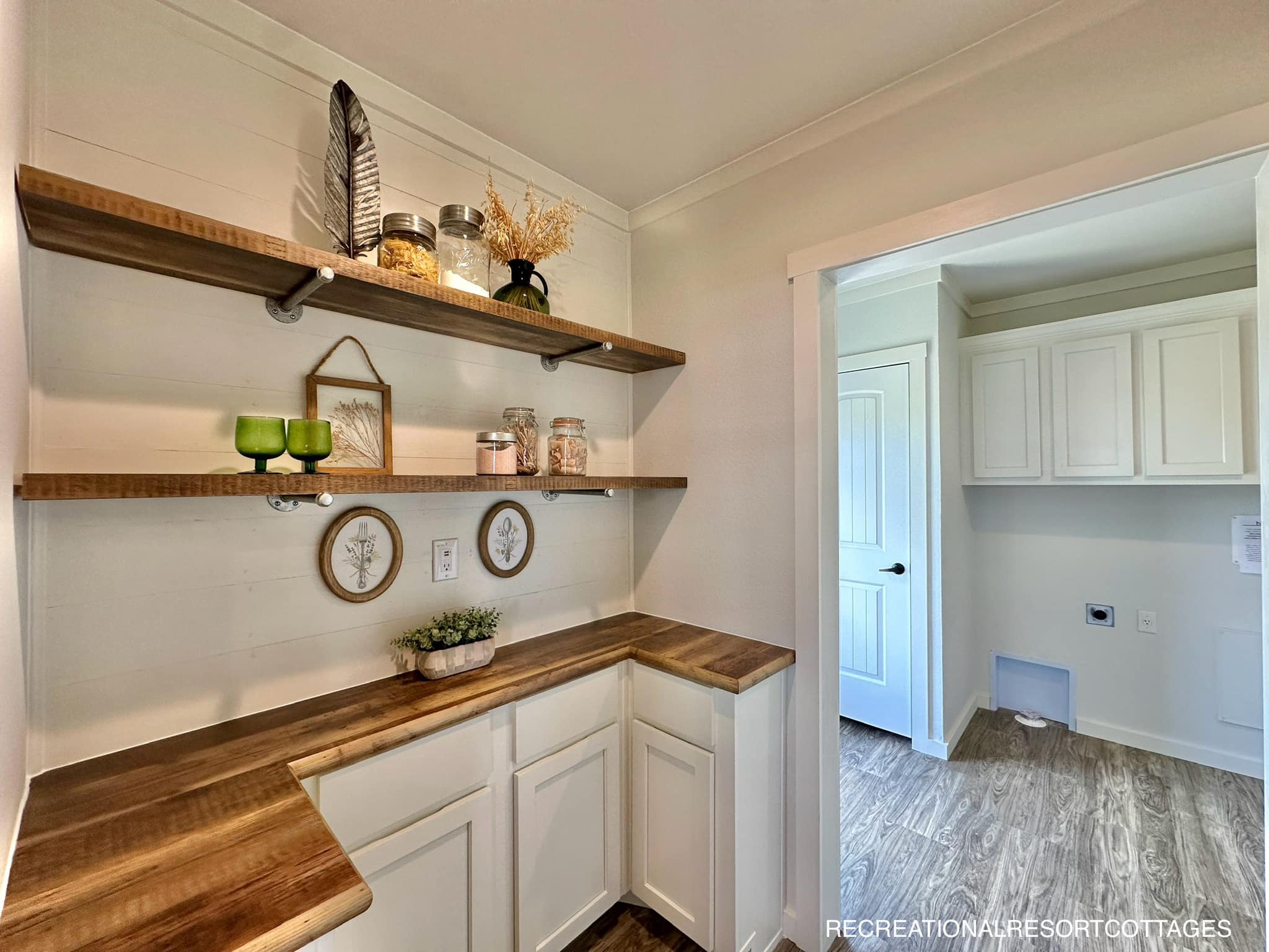 A cozy pantry with wooden shelves displaying jars and decor, like green glasses and framed art. White cabinets and rustic wood counters add charm.