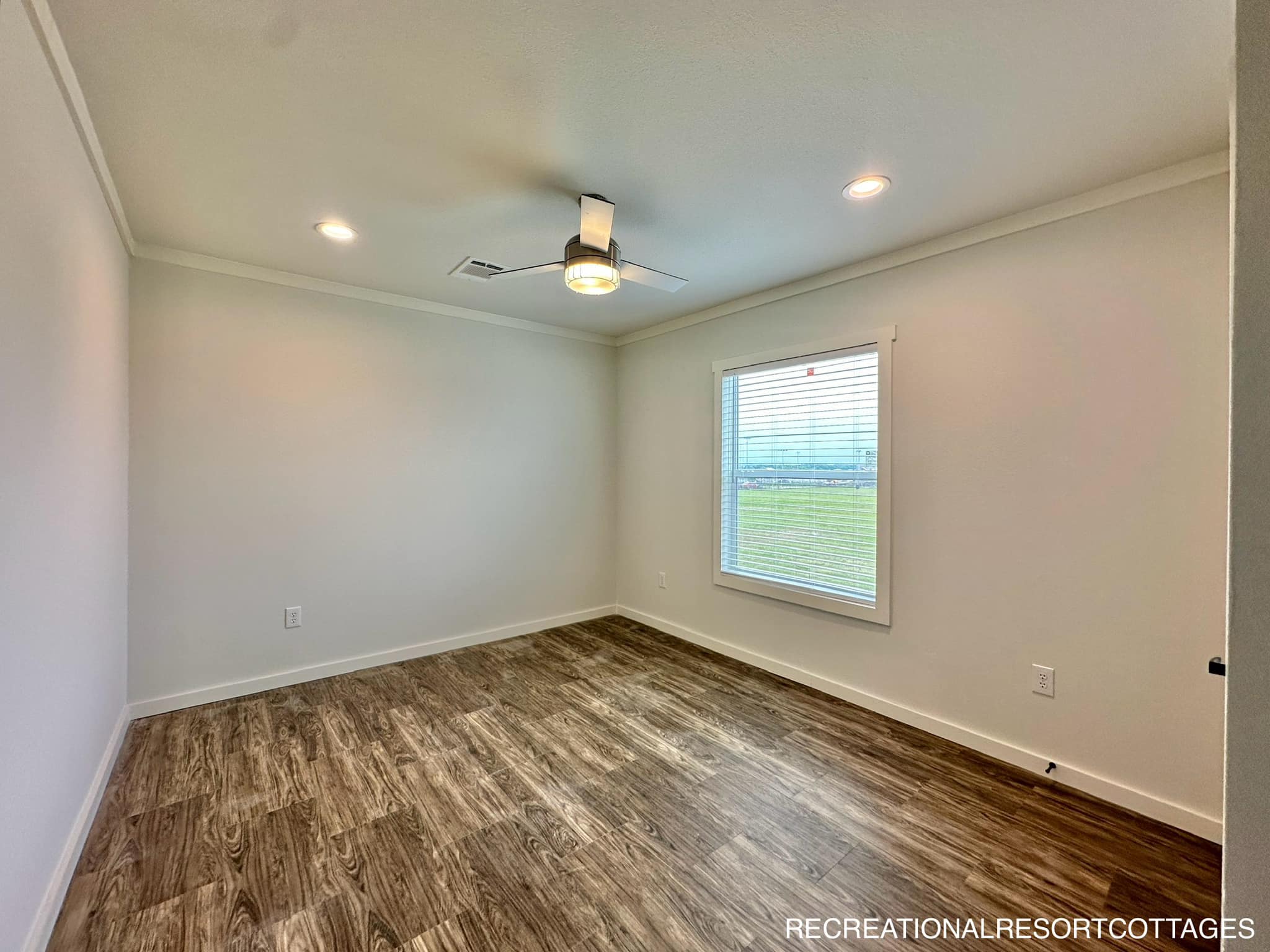 A minimalist, empty room with light wooden floors, white walls, a ceiling fan with a light, recessed lighting, and a window overlooking a green field.