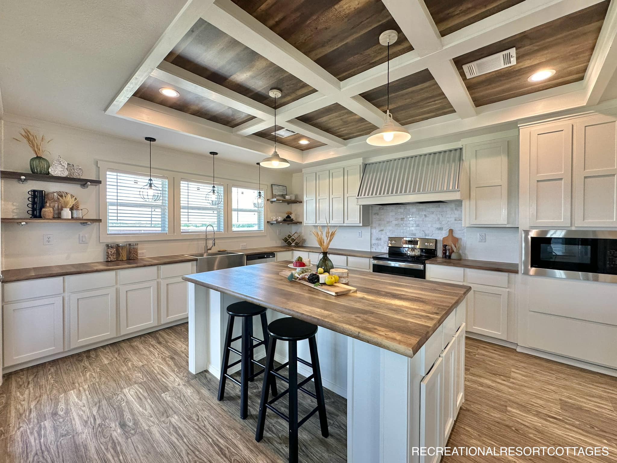 Spacious kitchen with white cabinets, wood countertops, and a large island with two stools. Rustic decor, coffered ceiling, and hanging lights.