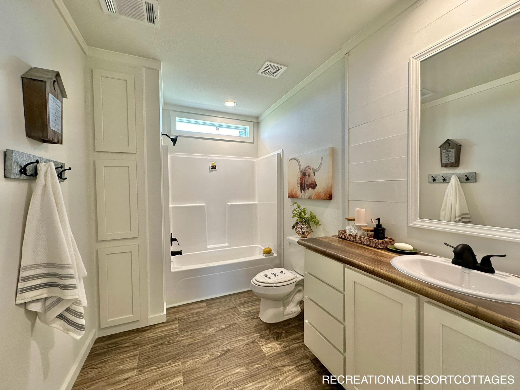 Modern bathroom with wood-look flooring, white vanity, and black fixtures. A tub-shower combination is at the back, accentuated by a small window. Decor includes a plant and framed art. Cozy and inviting.