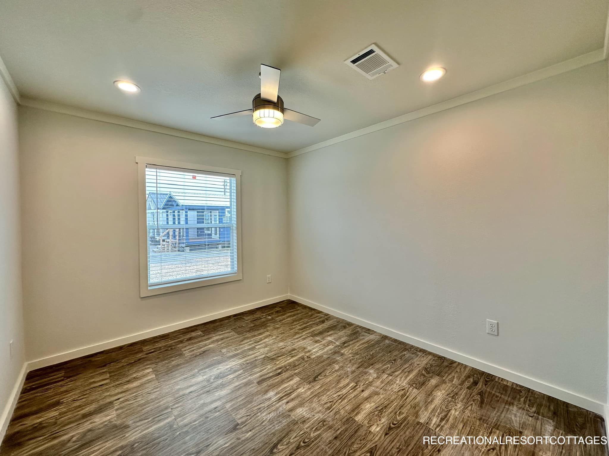 Empty room with wood flooring and beige walls, featuring a ceiling fan with light. A window shows a glimpse of houses outside. Bright and modern tone.