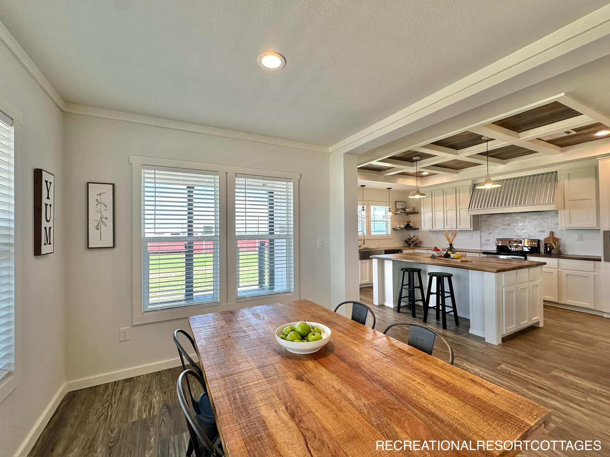 Modern kitchen and dining area with a rustic wooden table, bowl of green apples, and industrial chairs. Bright with natural light, wooden floors, white cabinets, and a coffered ceiling. Cozy and welcoming.