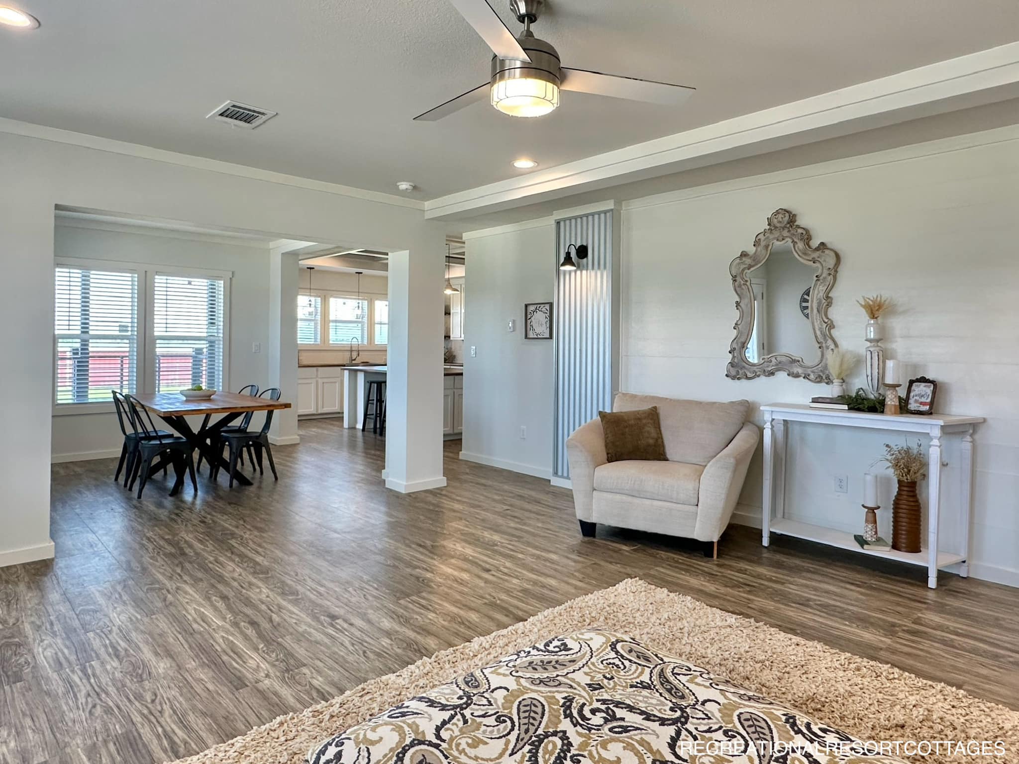 Bright, open living space with wooden floors, a beige armchair, decorative mirror, and wall art. Dining area and kitchen visible in background. Cozy, inviting feel.