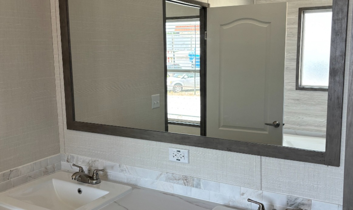 Modern bathroom with a large mirror above a white sink on a marble countertop. A half-open door and a window reflect in the mirror, creating a spacious feel.