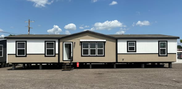 A beige and white manufactured home with a central front door, flanked by windows and set on a gravel lot under a bright blue sky with fluffy clouds.