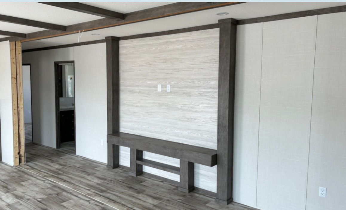 Empty living room with light wood flooring, a textured wood accent wall, and dark wood beams and trim. Visible doorway leads to a bathroom. Modern, minimalist tone.