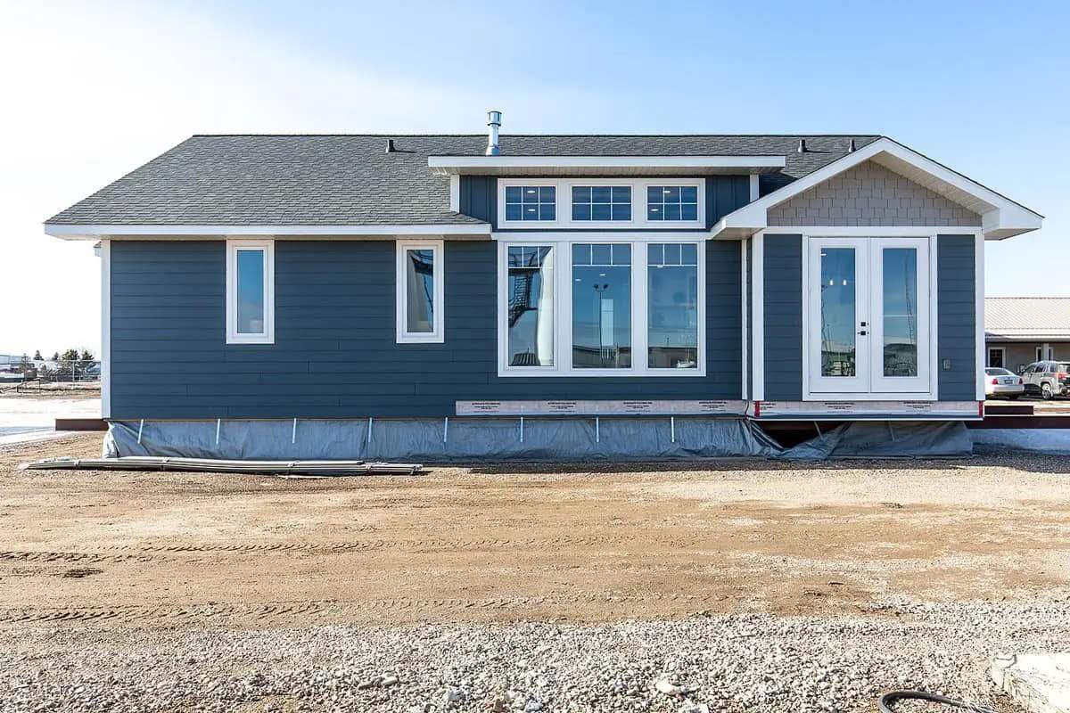 A single-story, blue modular home with white trim sits on a gravel lot. The home features large windows and a side door, under a clear blue sky.