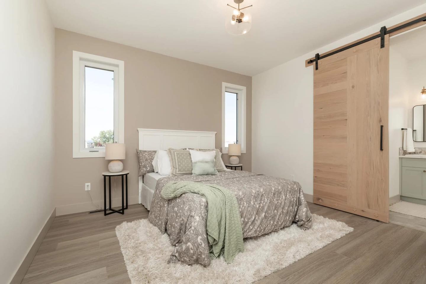 A cozy bedroom with a plush bed adorned with gray floral bedding and green accent pillows. Light wood sliding barn door leads to a bathroom. Minimalistic decor with warm lighting creates a calm, inviting atmosphere.