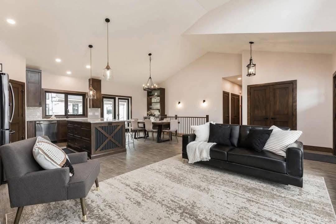 Spacious modern living area with a black sofa, gray armchair, and a large rug. Open-concept kitchen and dining area with pendant lighting and dark wood accents. Cozy, inviting atmosphere.