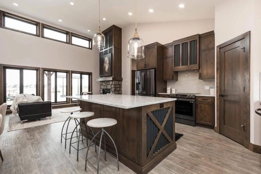 Spacious kitchen with dark wood cabinets, a large island with stools, pendant lights, and modern appliances. Open living area with large windows. Cozy and elegant.
