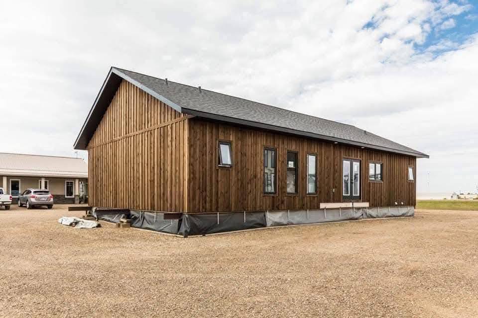 A rustic wooden cabin with a gray shingled roof stands on a gravel lot under a partly cloudy sky. There are parked cars and other buildings nearby.