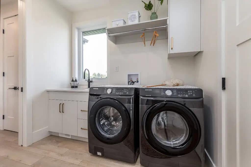 A modern laundry room with a sleek, black washer and dryer set beside a white countertop and sink. Shelves hold decor and hangers. Bright, clean ambiance.