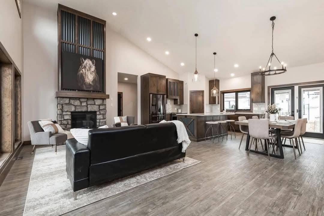 Spacious open-plan living room and kitchen with wooden floors. A cozy seating area with a black sofa faces a stone fireplace with a lion painting. Contemporary dining area and modern kitchen feature warm wooden tones, creating an inviting atmosphere.