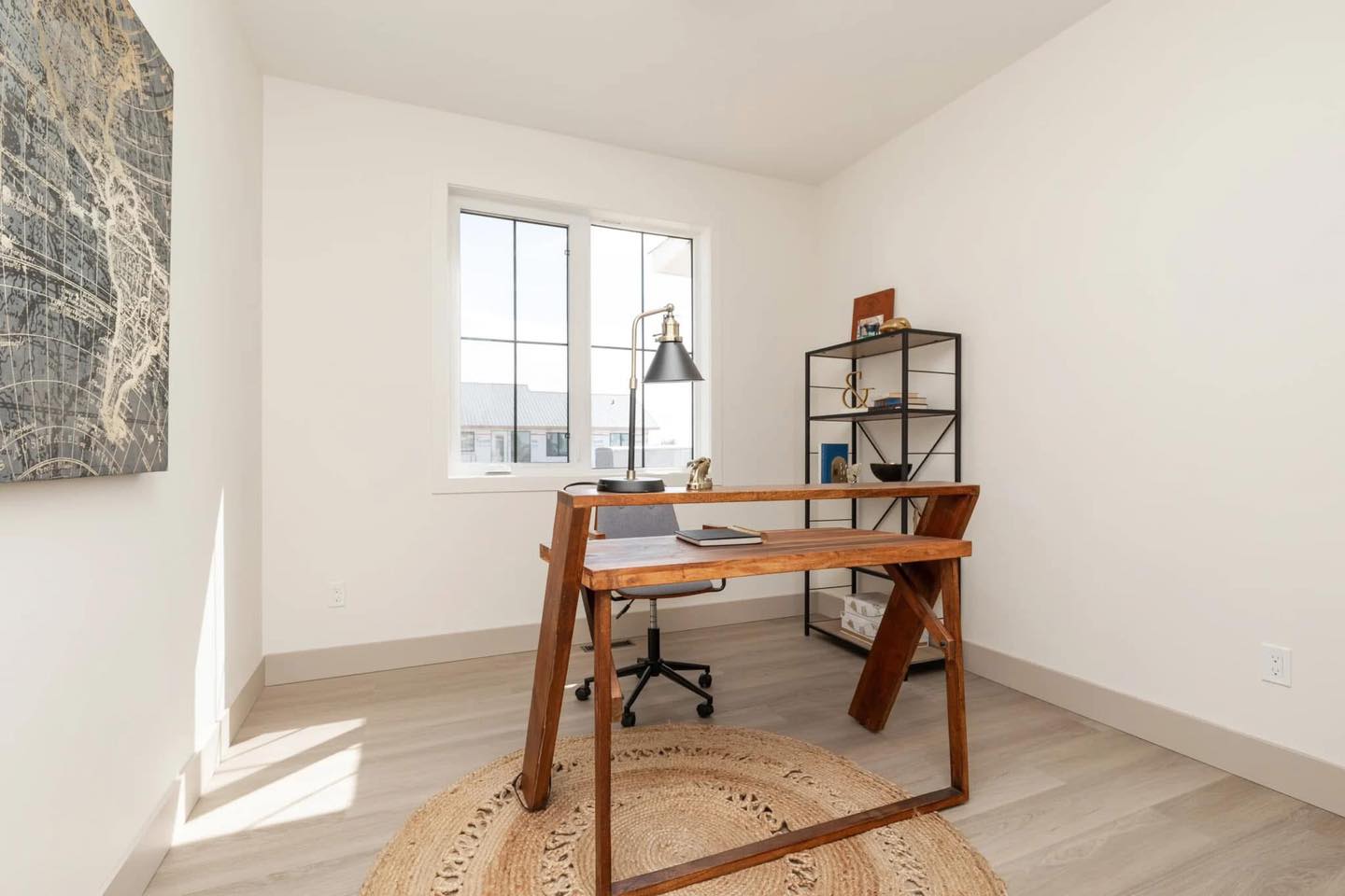 Bright minimalist home office with large window. A wooden desk and chair sit on a circular rug. A shelf with decor adds a modern touch.