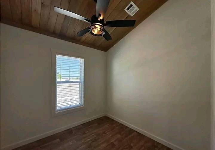 Small, empty room with a wood ceiling and dark ceiling fan above. A single window with blinds allows natural light, highlighting the warm wood floor.