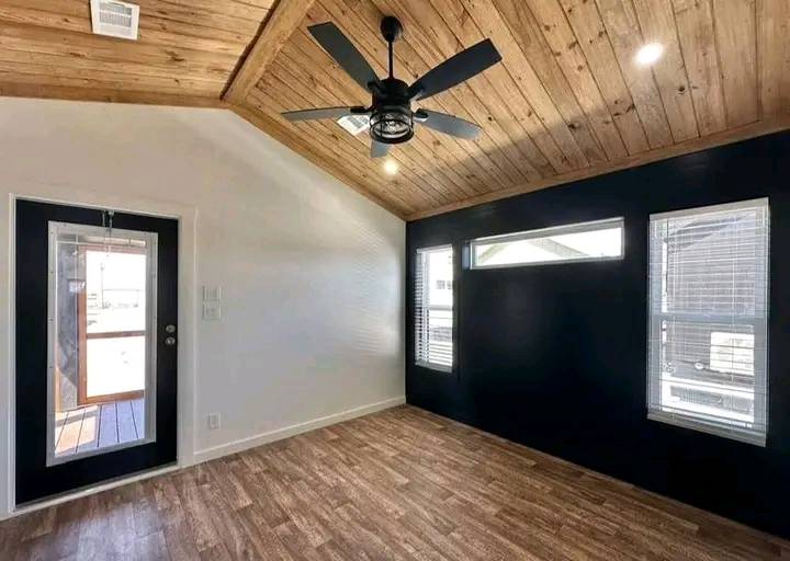 Alt text: "Modern room with wooden ceiling and floor, black accent wall, and three windows. A ceiling fan hangs from the vaulted ceiling, creating a cozy atmosphere."
