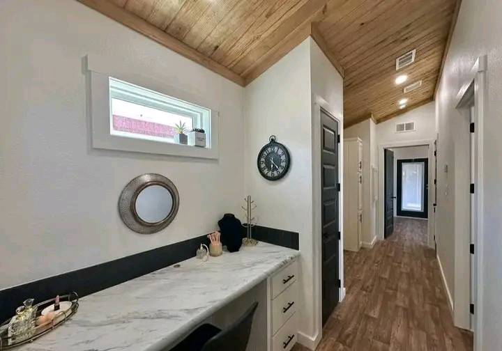 A modern hallway with a wooden ceiling and floor. On the left, a white marble desk with decor, a small window, and circular mirror. A black wall clock is on the right.